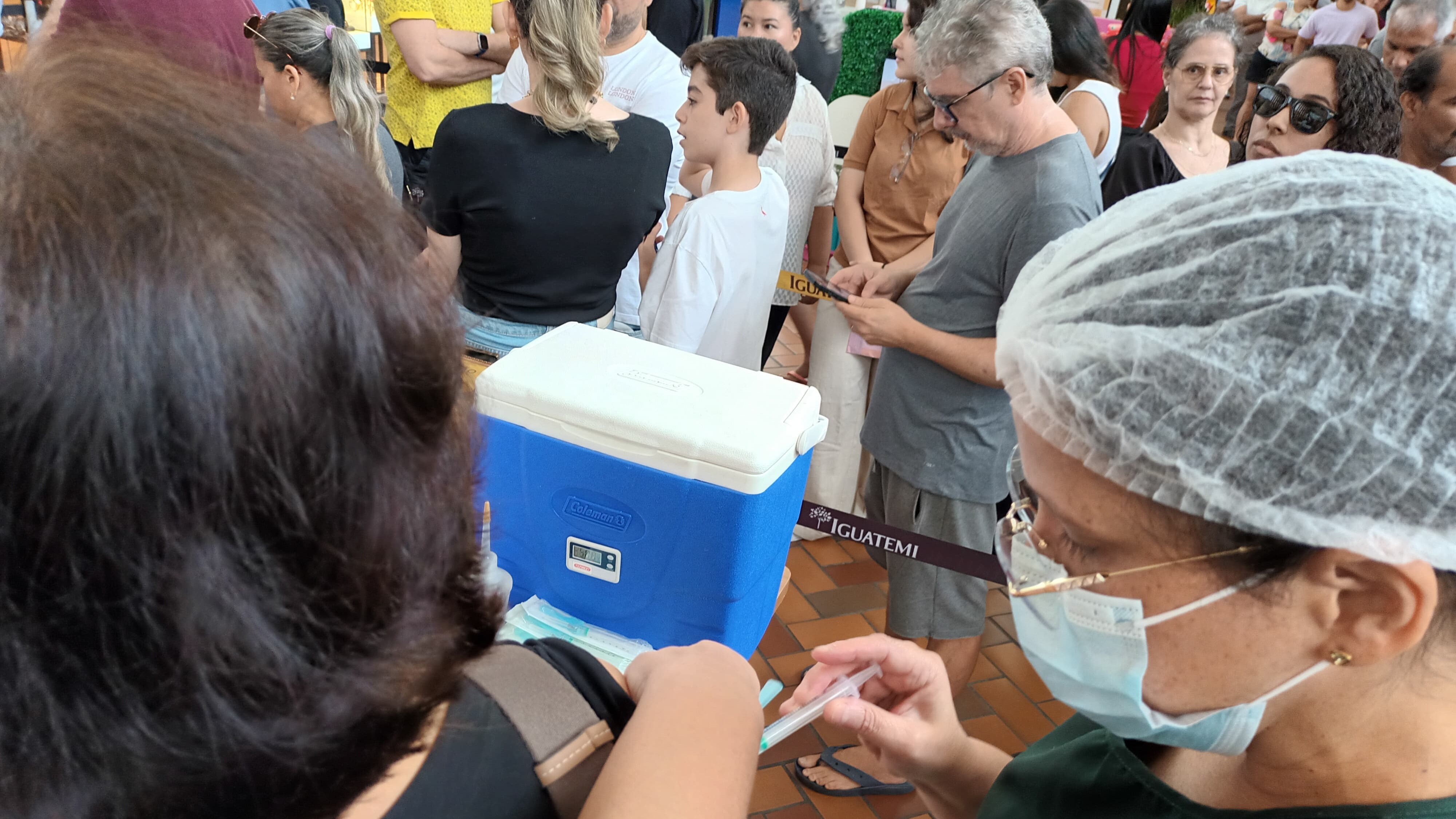 enfermeira vacina uma pessoa e, ao fundo, uma fila de pessoas aguarda